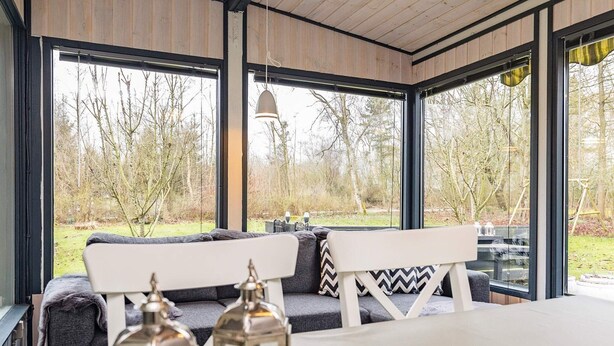 A covered outdoor seating area with a bench and table, large windows, and a view of trees. - Secluded Forest Retreat-by Traum (Roslev)