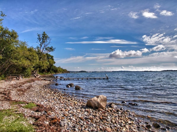 A pebbled beach with a wooden pier extending into calm waters, surrounded by trees and a clear sky.