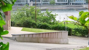 A modern building with large glass windows, a paved area with benches, and a body of water in the background.