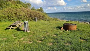 A grassy area with a picnic table, a fire pit, and a trash can near a body of water.