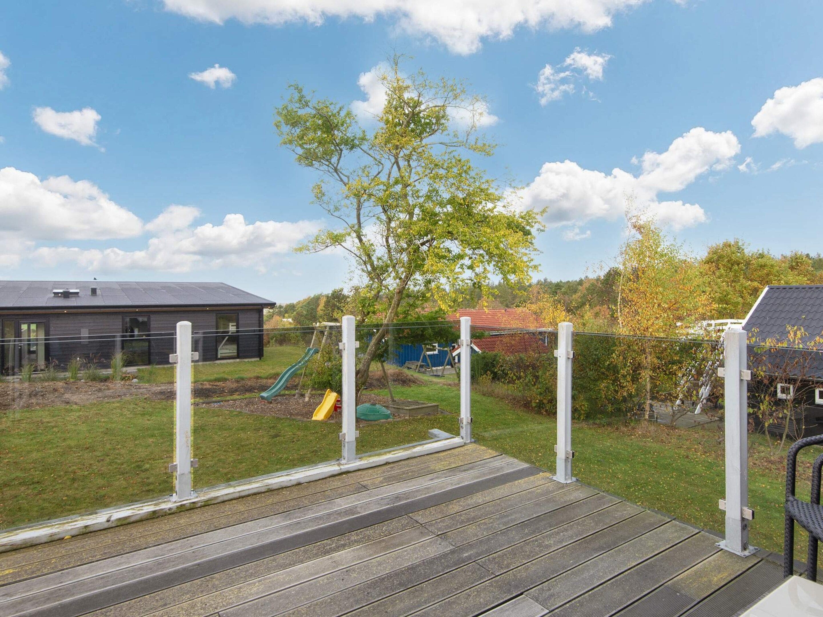 Una terraza de madera con un parque infantil, que incluye toboganes y columpios, rodeada por una zona vallada y una casa al fondo.