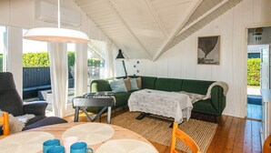 Ein gemütliches Wohnzimmer mit einem grünen Sofa, einem Esstisch mit Untersetzern und Tassen, einer Lampe und Blick ins Freie.