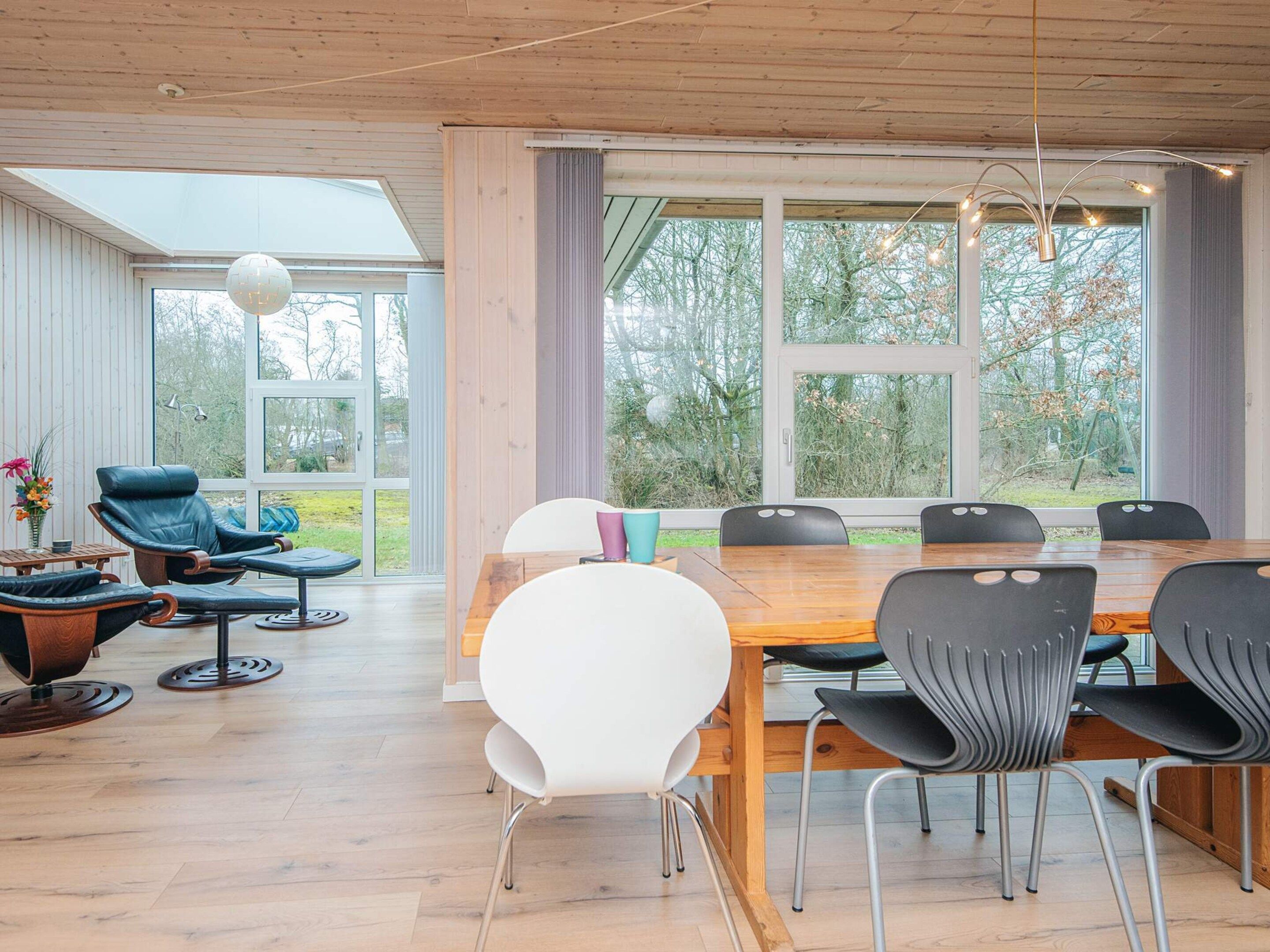 Een moderne eetruimte met een houten tafel en stoelen, een groot raam met gordijnen, en een zithoek met een leren fauteuil en een bijzettafeltje.