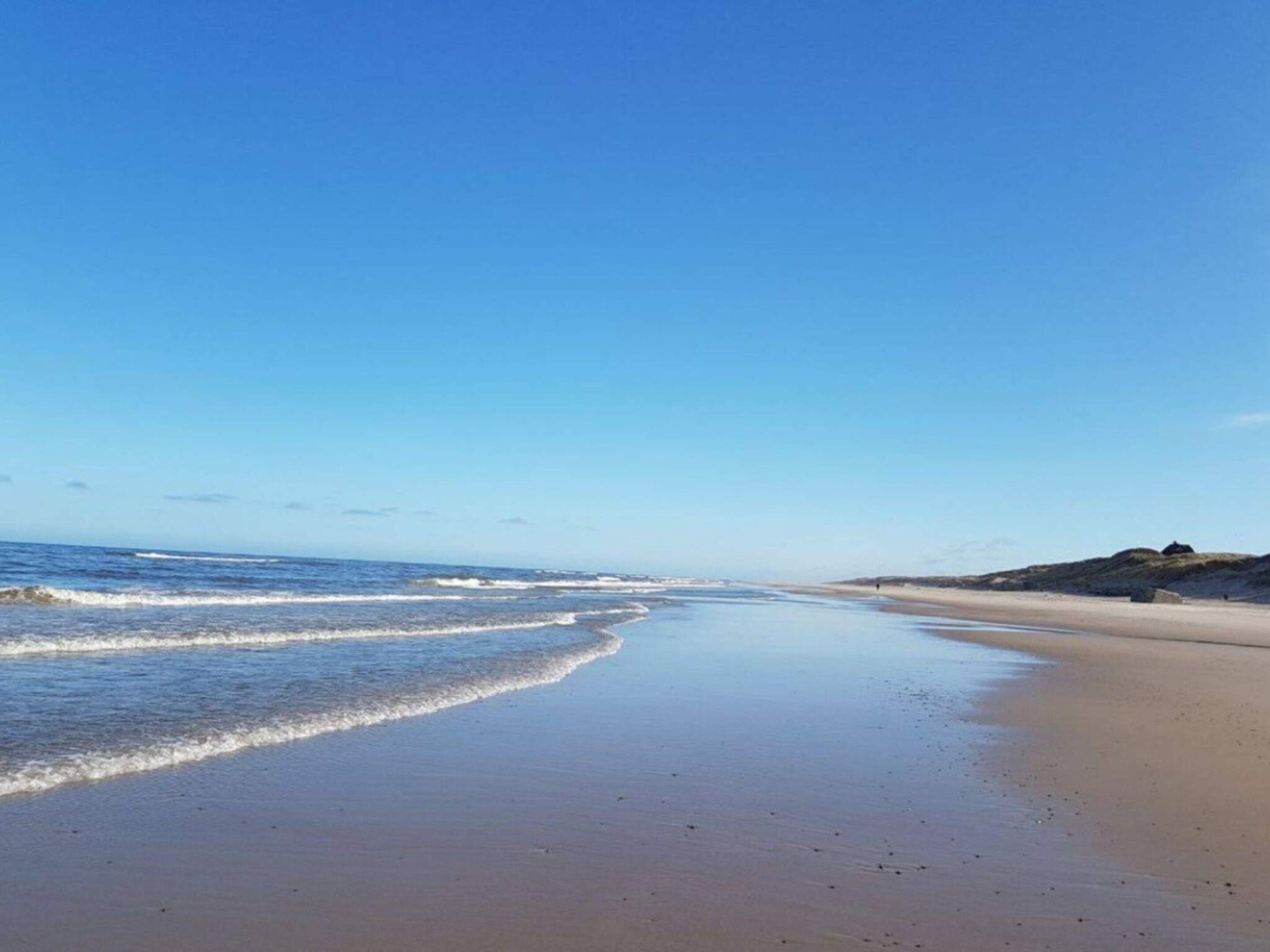 Une plage avec un ciel dégagé, des vagues calmes et une colline qui se dessine à l’horizon.
