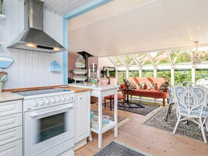 A kitchen with a stove, oven, and white cabinetry. There is a dining area with a table and chairs, and a sofa. The room has a large window and a ceiling fan. - Holiday Home in Snedsted near Sea (Thisted)