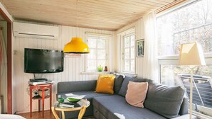 A cozy living room with a grey sofa, a television, a yellow pendant light, and a window with a view of trees. - Seaside Charm Near Blokhus Town-by Traum (Blokhus)