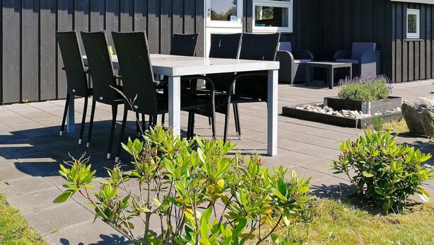 A patio with a table and chairs, a small garden area, and a building with a dark exterior and a sloped roof. - 6 Person Holiday Home in Humble (Sydlangeland)
