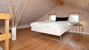A bedroom with a sloped ceiling, wooden walls, a bed with white bedding, a small table with a clock, and a vase with branches. - 6 Person Holiday Home in Humble (Sydlangeland)