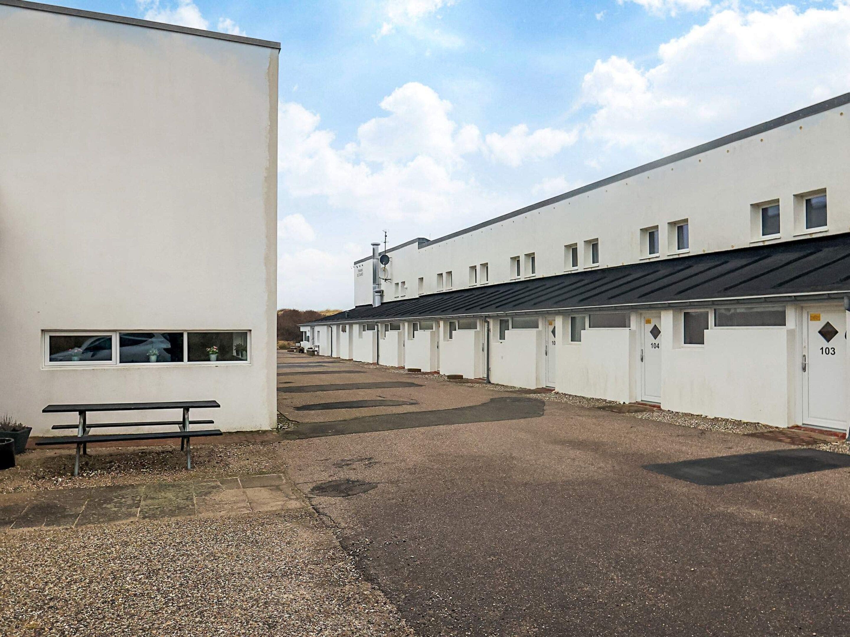 A row of white buildings with numbered doors, a gravel path, and a bench.