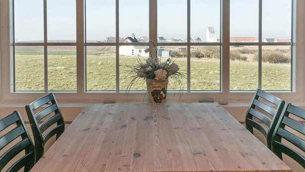 A dining area with a wooden table and chairs, a vase with flowers, and a large window with a view of a grassy field and buildings. - 12 Person Holiday Home in Lemvig (Lemvig)
