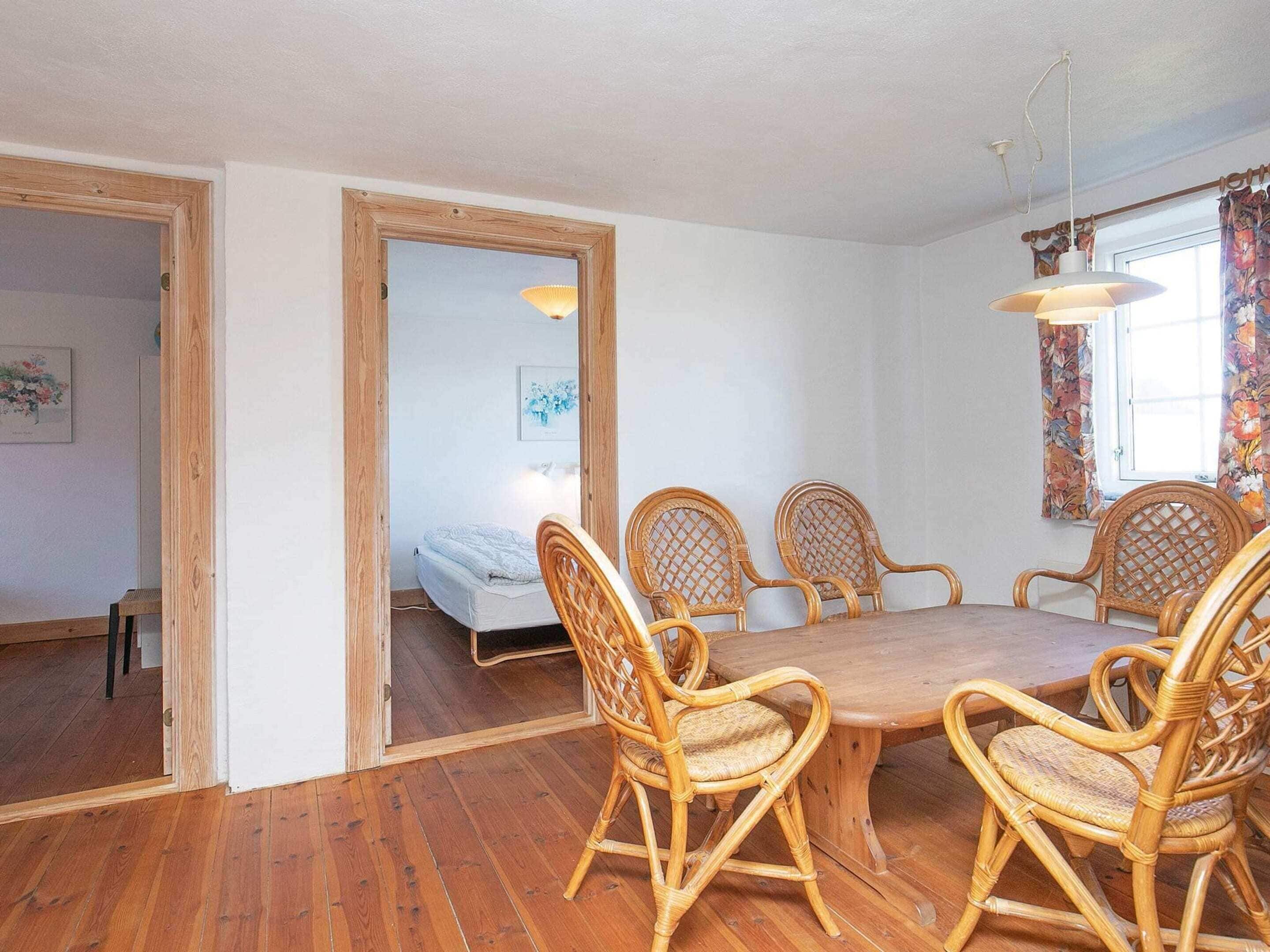 A room with a wooden dining table surrounded by wicker chairs, a bed with a white mattress, and a window with curtains.