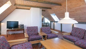 A cozy attic room with wooden beams, a sloped ceiling, and a skylight. There are two wooden tables, several patterned chairs, and a television on a wooden stand. - 12 Person Holiday Home in Lemvig (Lemvig)