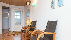 A room with wooden flooring, two wicker chairs, a small table, a wall clock, and framed artwork. - Holiday Home in GrenÃ¥ near Sea (Grenaa)