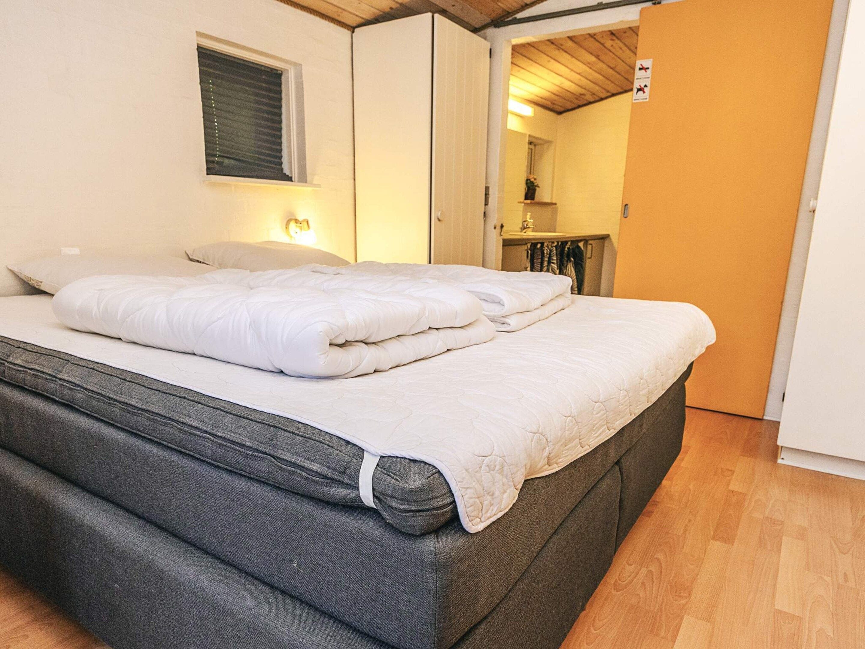 Un letto con materasso grigio e biancheria da letto bianca, una finestra con persiane e un soffitto in legno.