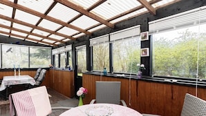 A covered patio with wooden walls, a round table with a pink tablecloth, wicker chairs, and a view of trees through large windows. - 5 Person Holiday Home in Gedser (Gedser)