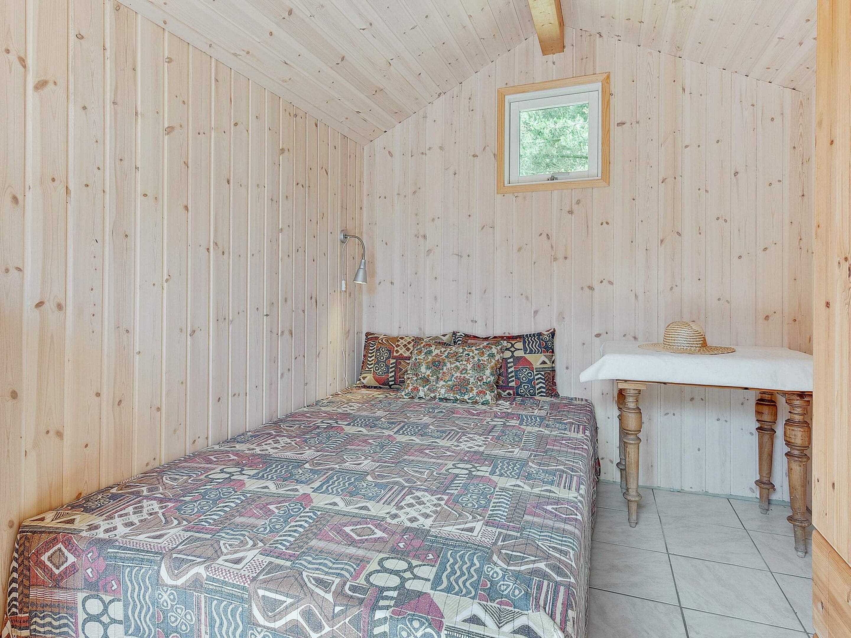 Une pièce avec un plafond en lambris de bois, un lit avec une literie à motifs, une petite table de chevet avec une lampe et une fenêtre donnant sur des arbres.