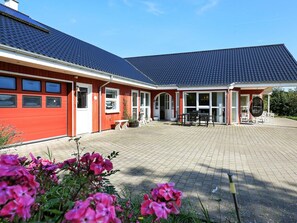 A red building with a tiled roof, a covered patio with outdoor furniture, and a garden with pink flowers.
