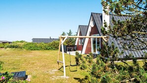 A modern house with a sloped roof, a swing set, and a grassy yard with pine trees. - 6 Person Holiday Home in Hvide Sande (Hvide Sande)
