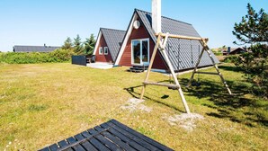 A red house with a sloped roof and a chimney, a wooden swing set, and a picnic table in a grassy yard. - 6 Person Holiday Home in Hvide Sande (Hvide Sande)