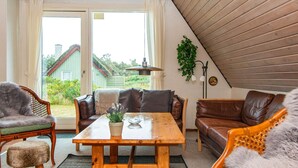A cozy living room with a wooden table, leather sofas, and a view of a green house through large windows. - 6 Person Holiday Home in Hvide Sande (Hvide Sande)