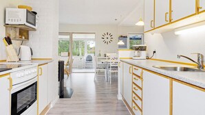 A modern kitchen with white cabinetry, a built-in oven, a microwave, and a sink. There is a dining area with a table and chairs visible through an open door. - 6 Person Holiday Home in Saeby (Sæby)