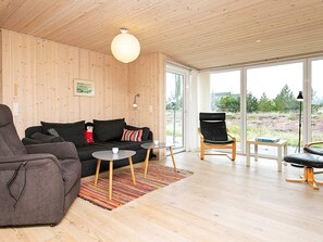 A modern living room with a sofa, armchair, and coffee table, wooden flooring, and large windows offering a view of the outdoors.