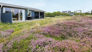 Une maison moderne dotée d’un patio couvert, d’une balançoire et d’un champ de fleurs violettes.
