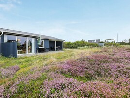 Et moderne hus med en overdækket terrasse, en gyngestativ og et felt med lilla blomster.
