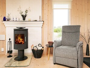 A cozy living room with a wood-burning stove, a grey armchair, and a window with a view of greenery.
