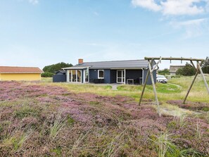 Et hus med en overdækket terrasse, gyngestativ og en bil parkeret i nærheden.