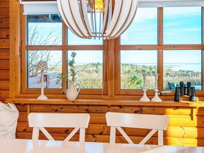 A cabin interior with wooden walls, two white chairs, a table, a lamp, a vase, and a view of the outdoors through large windows.