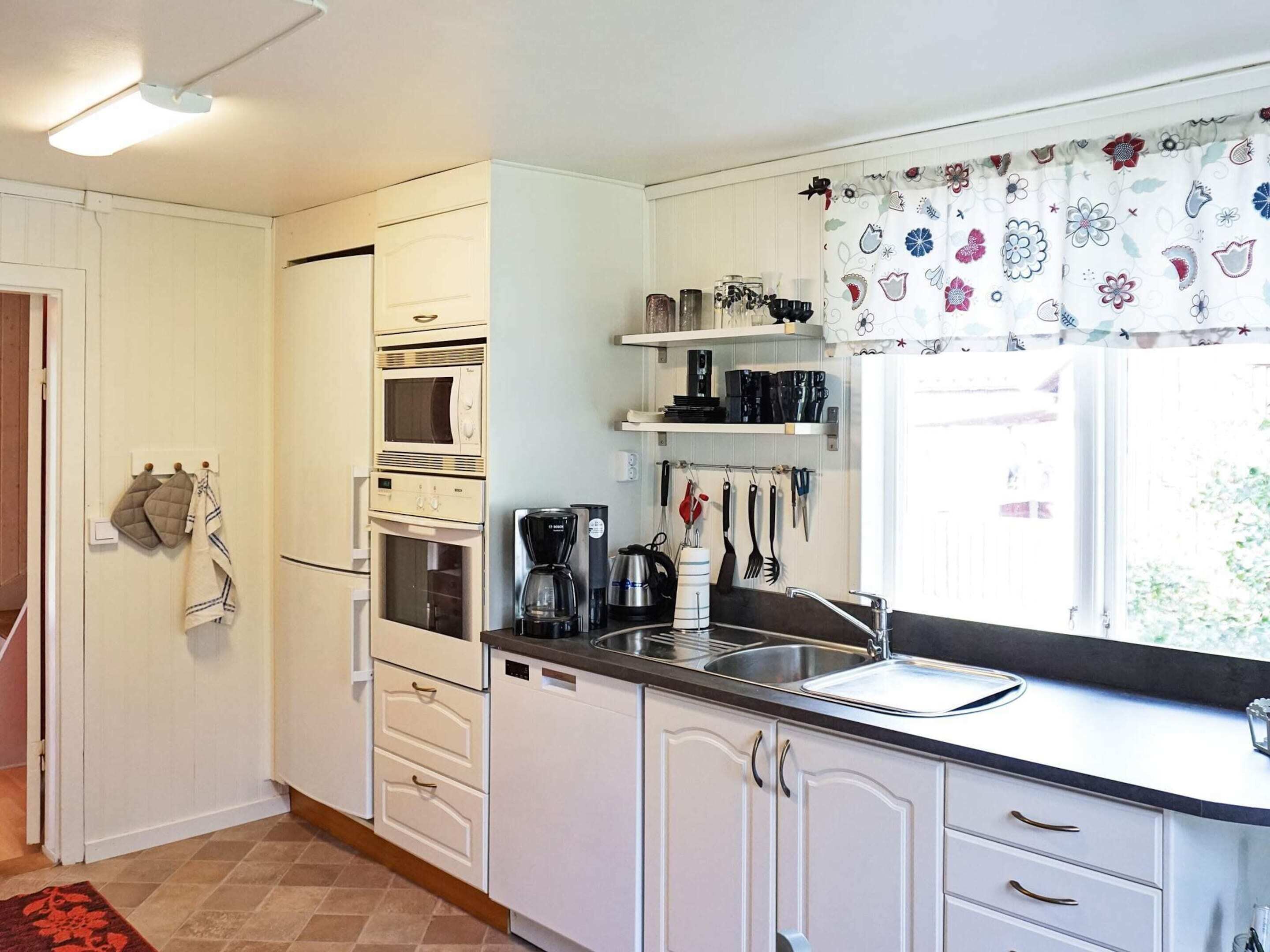 Una cucina compatta con mobili bianchi, forno integrato, macchina da caffè e finestra con tende a motivi.