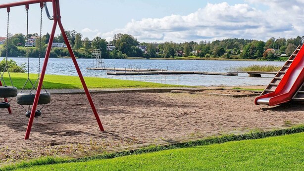 A playground with swings and a slide, a sandy area, and a lake with houses in the background.