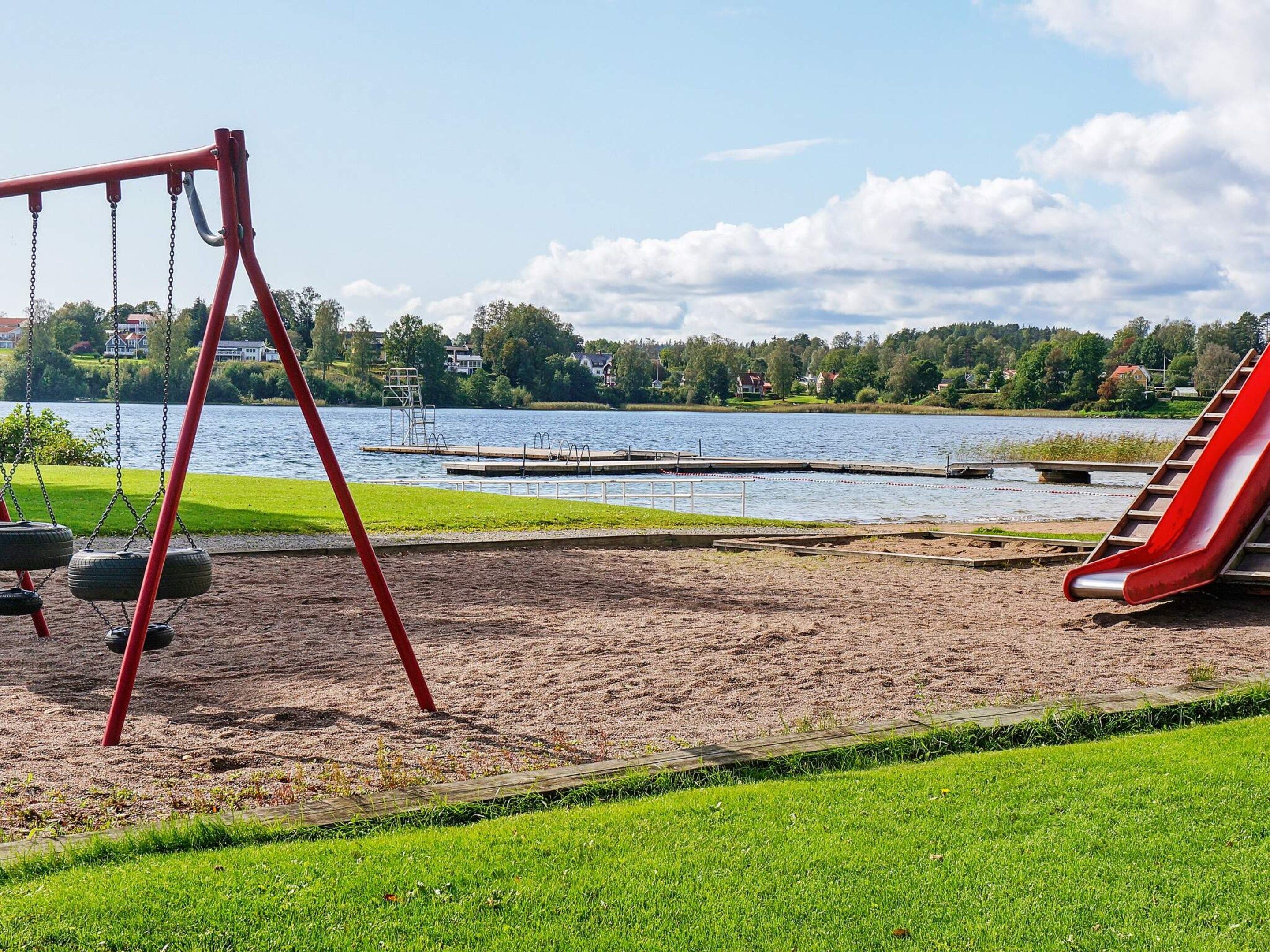 A playground with swings and a slide, a sandy area, and a lake with houses in the background.