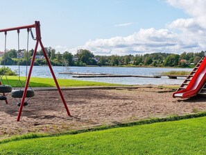 A playground with swings and a slide, a sandy area, and a lake with houses in the background. - 6 Person Holiday Home in Ed (Ed)