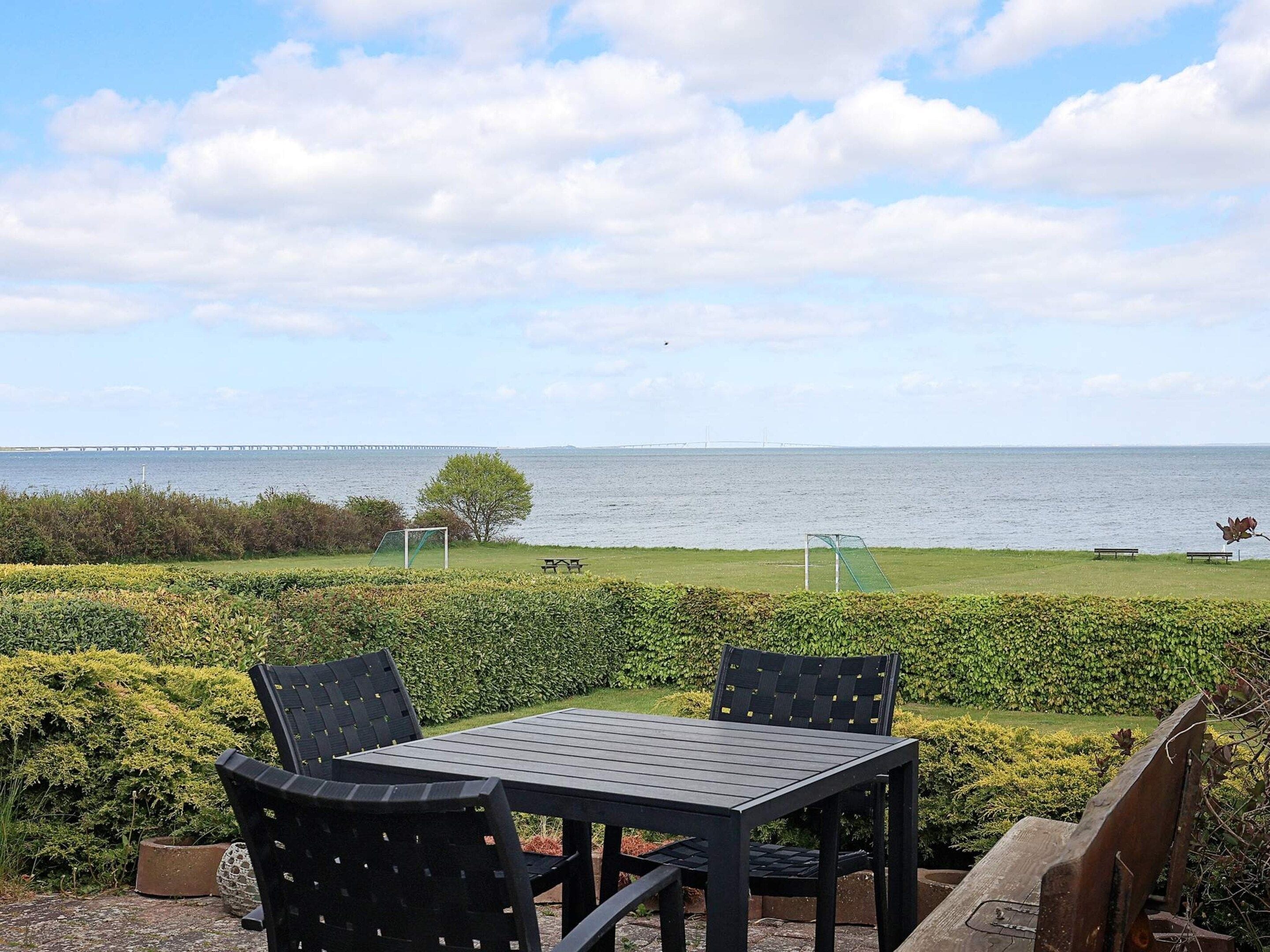 An outdoor seating area with a table and chairs, a view of the sea, and a distant bridge.