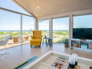 A modern living room with large windows, a yellow armchair, a glass coffee table, and a television. - 6 Person Holiday Home in Lokken (Lokken)