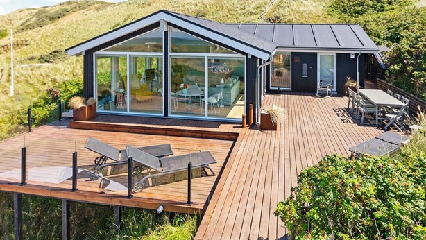 A modern house with a large glass facade, a wooden deck, and a view of the surrounding hills. - 6 Person Holiday Home in Lokken (Lokken)