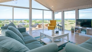 A spacious living room with a large sofa, a coffee table, and a television. The room features large windows offering a view of the beach and ocean. - 6 Person Holiday Home in Lokken (Lokken)