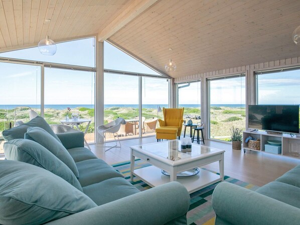 A spacious living room with a large sofa, a coffee table, and a television. The room features large windows offering a view of the beach and ocean. - 6 Person Holiday Home in Lokken (Lokken)
