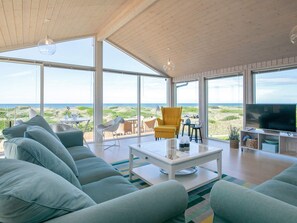 A modern living room with a large sofa, a coffee table, and a television. There are large windows offering a view of the beach and the ocean. - 6 Person Holiday Home in Lokken (Lokken)