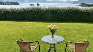 A round glass table with a vase of flowers, two wicker chairs, and a view of a lake with distant mountains.