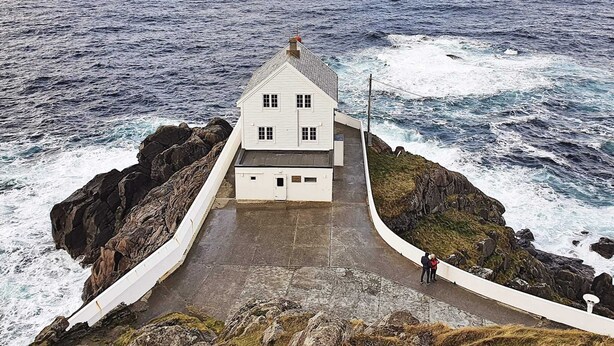A white building with a steep roof, located on a rocky coastline with the ocean in the background.