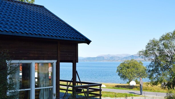 A wooden cabin with a large window, a balcony, and a view of a lake and mountains.