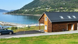 Ein Küstenhaus mit geneigtem Dach, einer gepflasterten Auffahrt und Blick auf Meer und Hügel im Hintergrund.