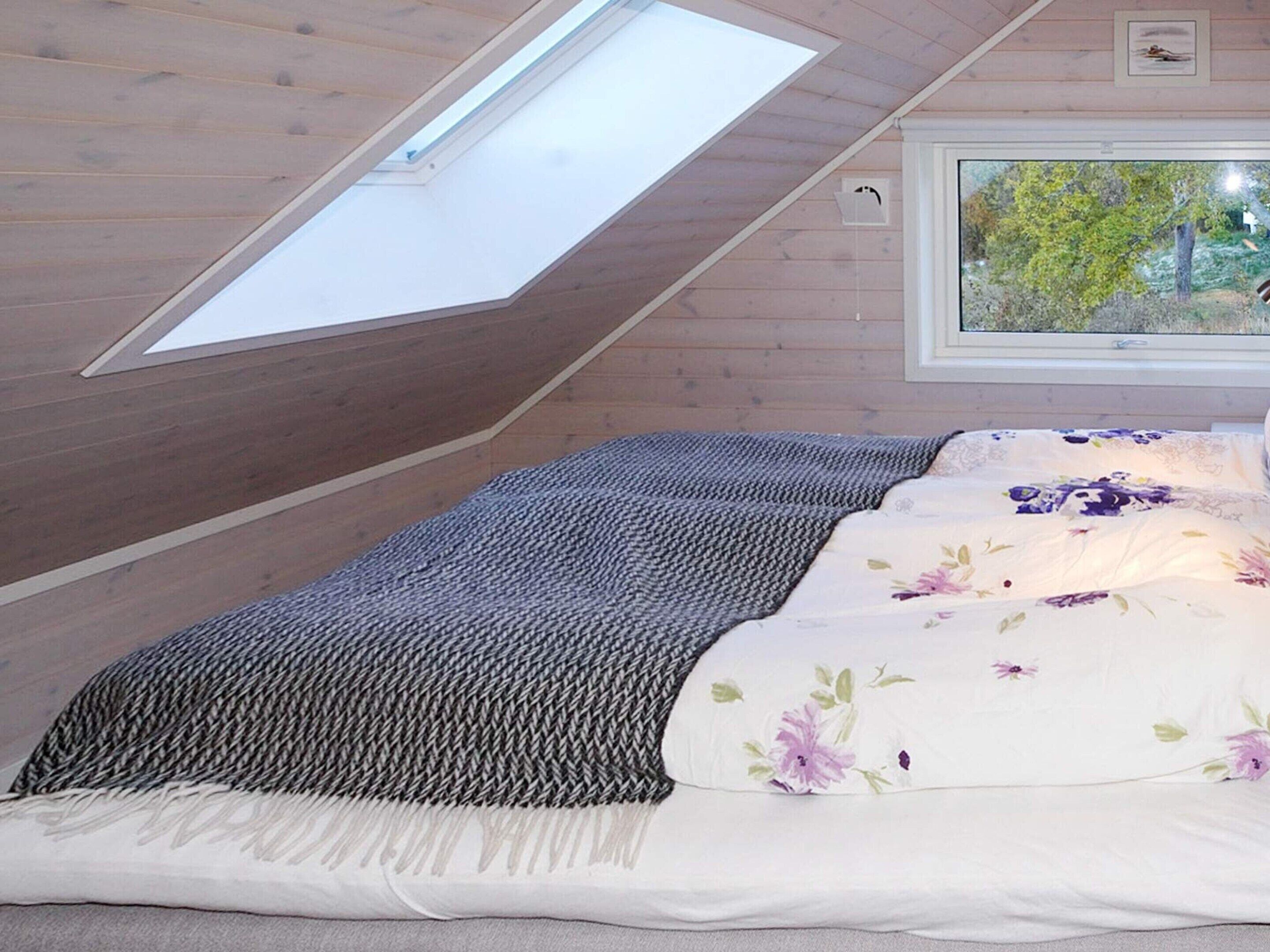 Ein Schlafzimmer mit Schräge und Dachfenster, ein Bett mit blumigem Überwurf und ein Fenster mit Blick auf Bäume.
