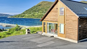 Ein modernes Holzhaus mit geneigtem Dach, großen Glasfenstern und einer gepflasterten Auffahrt, die zu einem Steg führt.