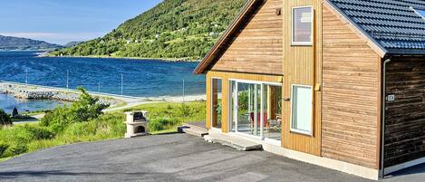 A modern wooden house with a sloped roof, large glass windows, and a paved driveway leading to a dock.