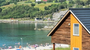 Eine Holzhütte mit Satteldach in der Nähe eines Sees, am Ufer befinden sich Personen.