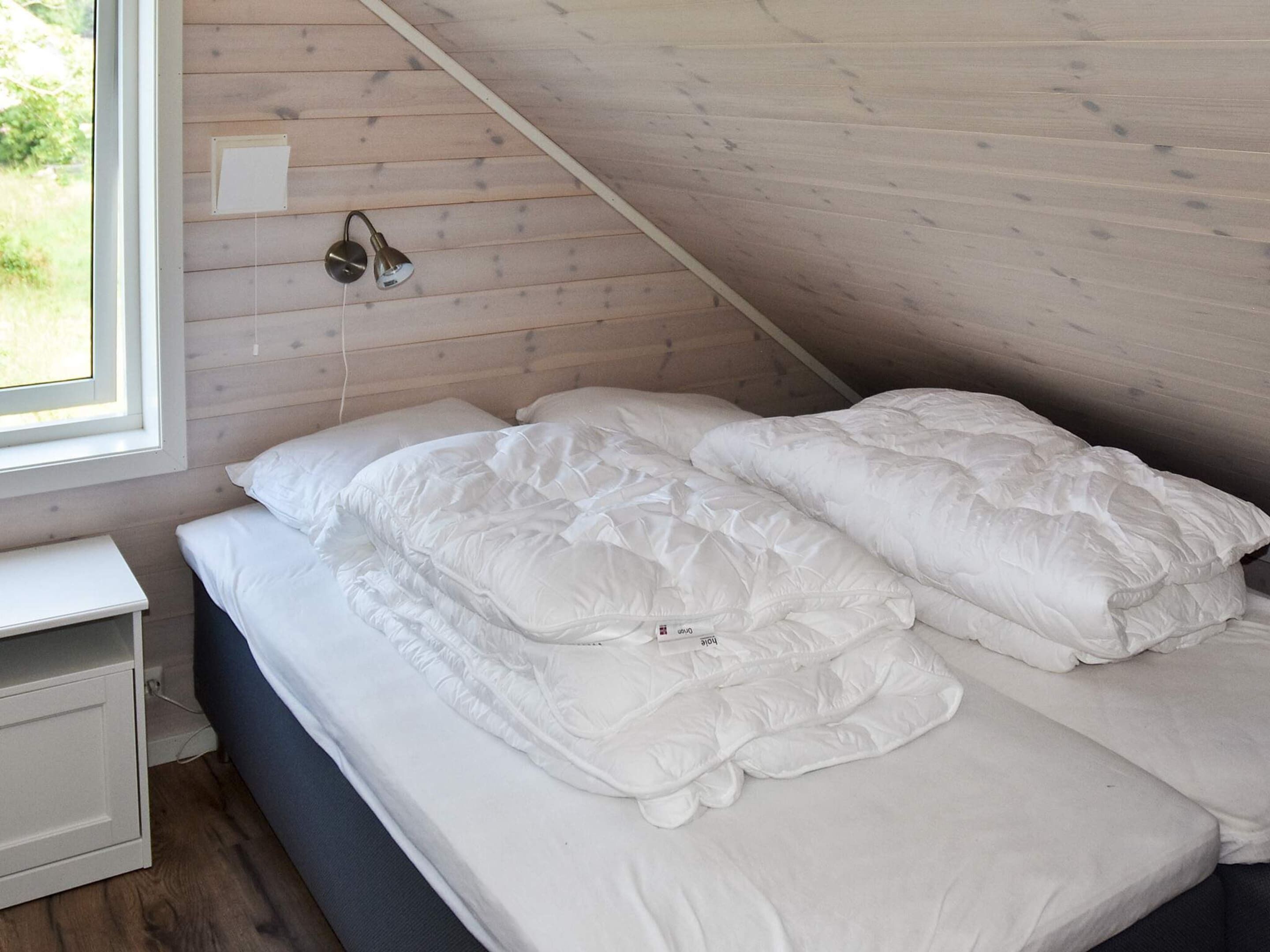 A bedroom with a sloped ceiling, a bed with white bedding, a bedside table, and a window with a view of greenery.